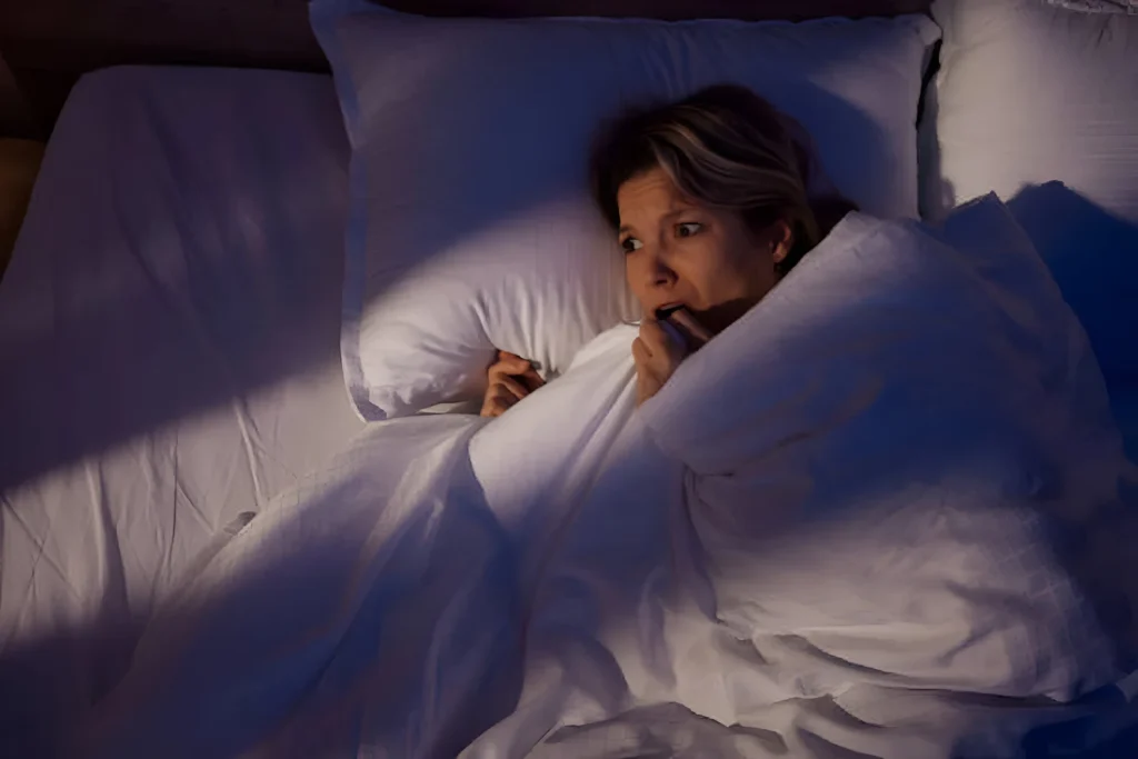 Persona en la cama con expresión de miedo o ansiedad, tapada con las sábanas en la oscuridad.